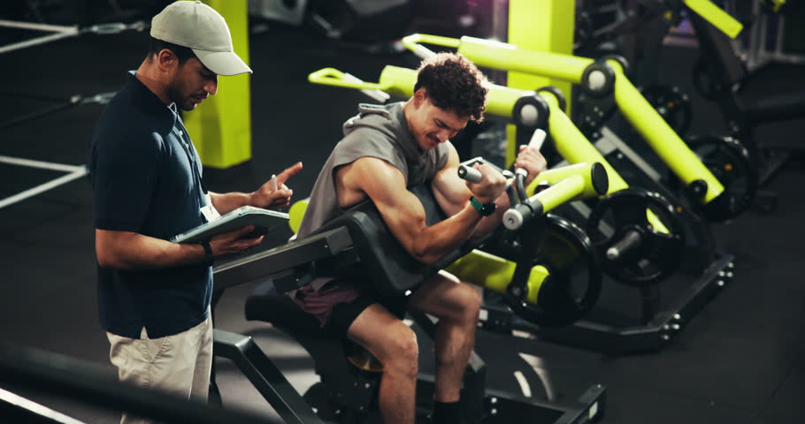 Men, personal trainer and bicep curl with machine for strength training, workout or exercise at gym. Male people, fitness instructor or coach writing with tablet for bodybuilding routine or checklist