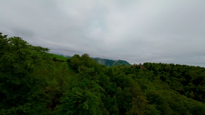 Aerial view of lush green forested mountains shrouded in mist and low-hanging clouds under a moody overcast sky, creating a serene and mysterious natural landscape.