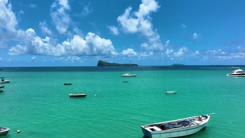 Aerial footage of a beachfront resort with tropical architecture and pools, facing a tranquil lagoon under a vibrant sunrise sky with boats and palm trees in Le Morne, Mauritius