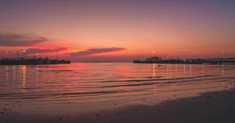 Colorful sky blending vibrant hues of orange, red, pink, and purple at sunset, with a calm sea reflecting a fishing harbor and industrial pier, creating a stunning seascape