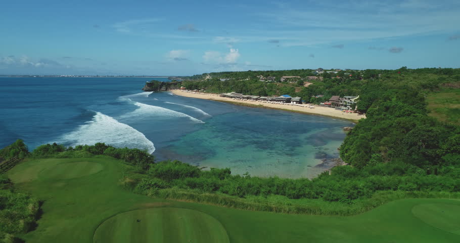 Lush green golf course sits adjacent to the sandy beach of Dreamland Beach in Bali, Indonesia, with waves rolling in from the ocean under a bright blue sky
