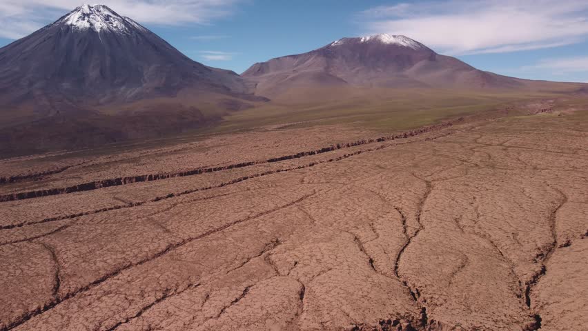 Aerial view of snow capped Licancabur Volcano at the Chile–Bolivia border. Cracked desert ground, volcanic slopes, and dramatic textures of the Andean plateau.