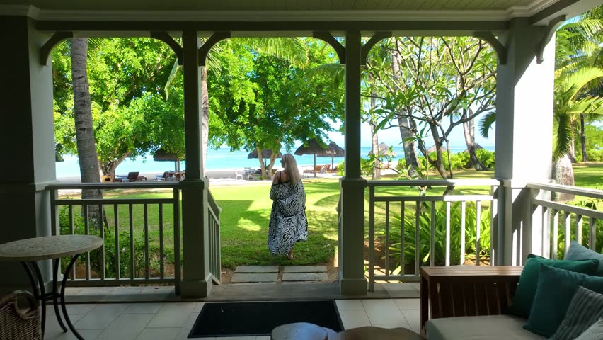 Aerial view of a blonde adult woman in a patterned robe walking from a tropical beach villa toward the sea in Mauritius