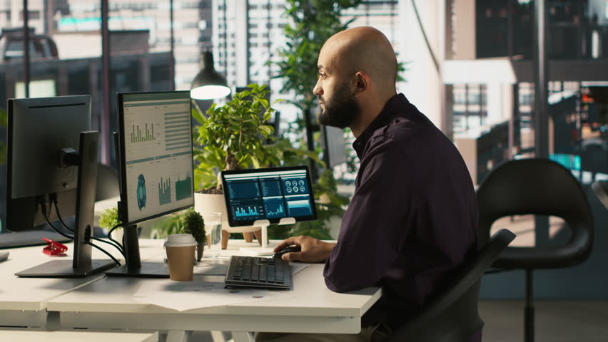 Arab office clerk working to hit key performance targets for revenue, profitability and operational efficiency. Middle Eastern employee at PC desk doing research analysis, camera A