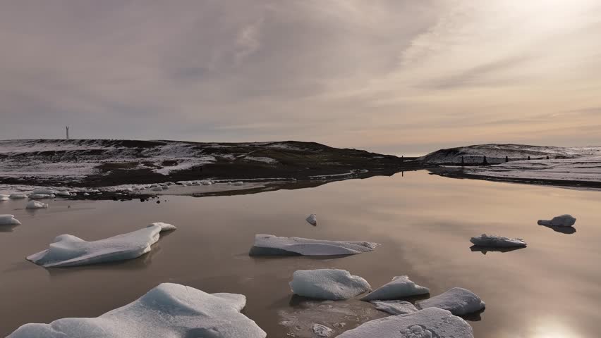 Iceland’s Fjallsárlón Glacier lake - smooth drone flight during golden hour with peaceful icy reflections, soft sunset sky and tranquil winter landscape.