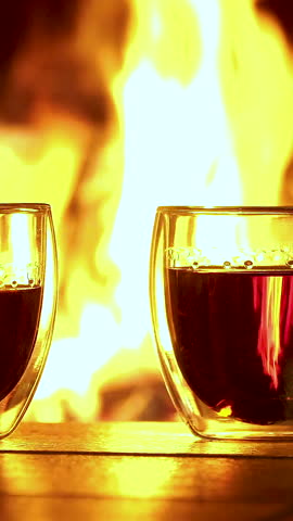 Steaming tea in double-walled glasses glows warmly by the blazing fireplace. Vertical footage