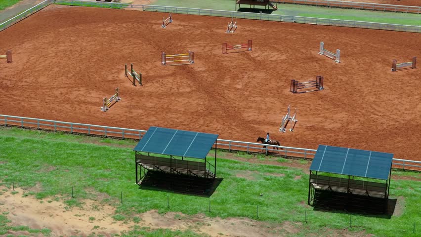 Training session at equestrian facility with horse jumping obstacles on red sand arena.