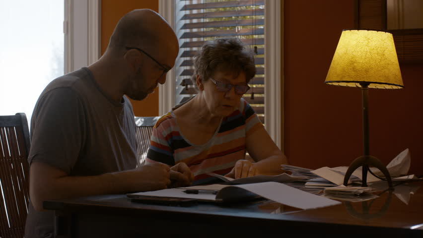 A middle-aged man and a senior woman sit together at a dining table in warmly lit home, sorting through paperwork. Woman dictates information, while man jots down notes in a notepad, with a back pen.