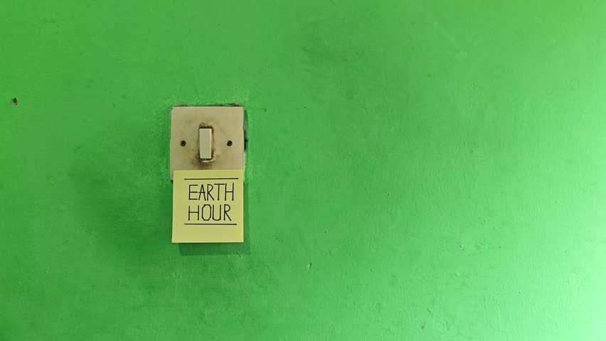A hand turning off a light switch with an Earth Hour sticky note on a green wall, promoting energy saving and climate action awareness
