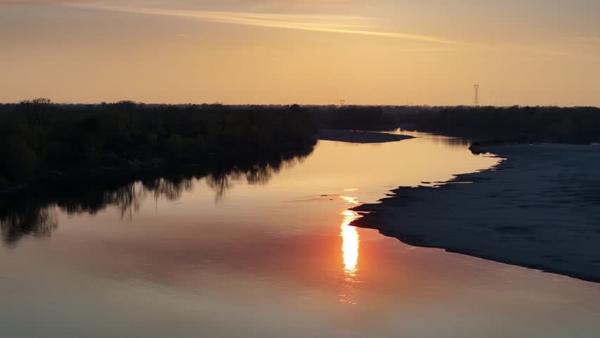 Warm sunlight reflects on the calm torrent of Adda waters of the Po River as the camera rises near Isola Serafini, showing riparian vegetation and sandy banks, aerial view, real time