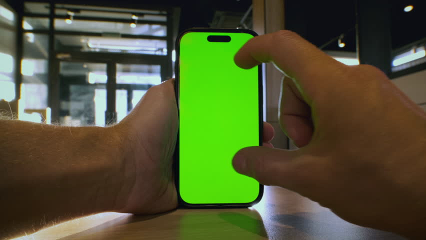 POV: Man holds a smart phone in his hands with a green screen for chroma key in cafe, restaurant. Male using gadget with app for delivery. Food court. Guy zoom out map on the phone display