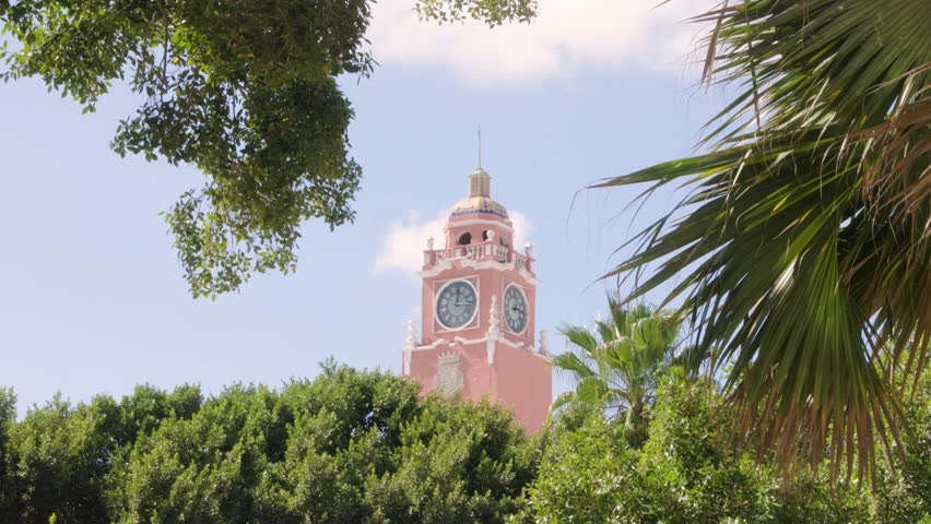 Picturesque view of the Palacio Municipal’s pink clock tower in Mérida, Mexico, framed by tropical palm leaves. The colonial architecture, pastel tones evoke the charm of this Yucatán capital
