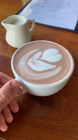 Food vlogger or travel influencer showing his or her coffee cup with flower or leaf drawing on table. Holding mug in restaurant or cafe. Enjoying delicious tasty coffee on sunny day POV shot ugc style