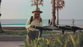 Blond female tourist wearing sunglasses and hat using smartphone while renting electric scooter near seaside promenade, enjoying urban mobility and vacation leisure - Powered by Shutterstock - Get 15% off with code: PIKWIZARD15