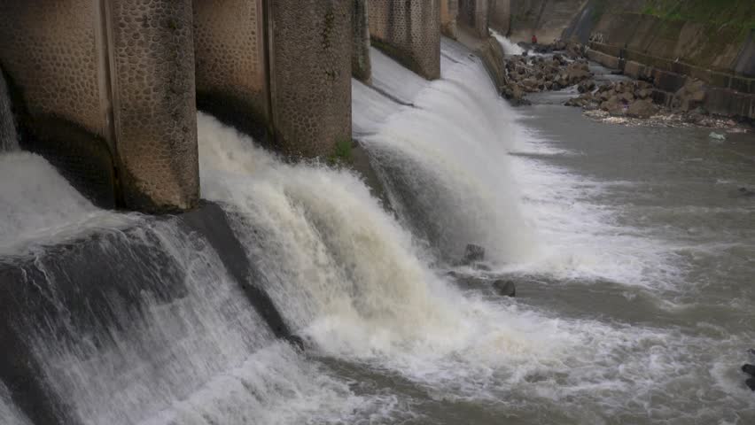 Multiple streams of water discharge forcefully from a large concrete dam, flowing into the river. Water management infrastructure.