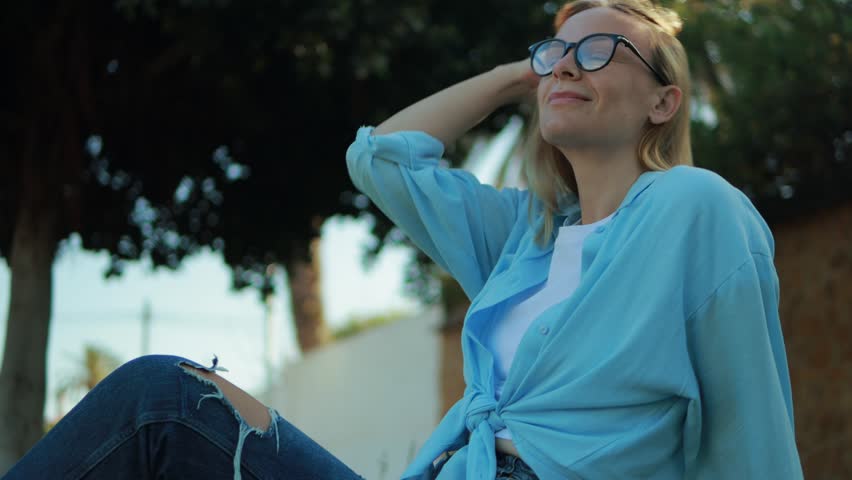 Blond woman in blue shirt, ripped jeans relaxing outdoors, smiling softly, touching hair under warm sunlight with carefree demeanor