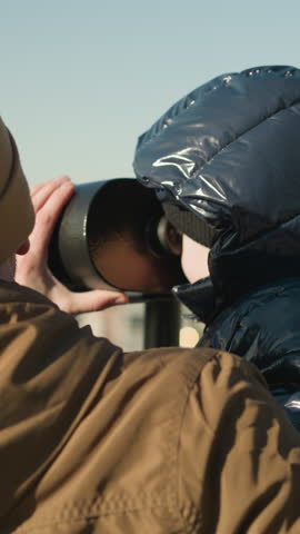 A father holds his son up to a public telescope, guiding him as the looks through it, exploring the distant cityscape, the father ensures a steady view