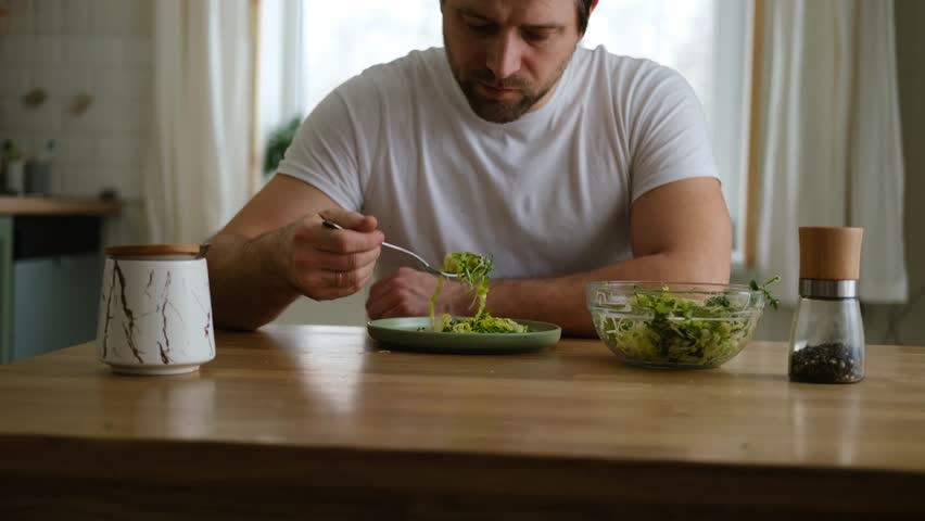 Handsome bearded man is eating green fresh vegan salad in the kitchen at home. Male sitting at table and eats vegetables, lettuce vegetarian meal. Happy athlete male enjoying Tasty healthy diet food.