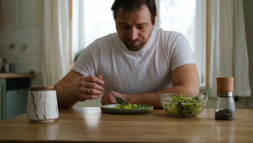 Handsome bearded man is eating green fresh vegan salad in the kitchen at home. Male sitting at table and eats vegetables, lettuce vegetarian meal. Happy athlete male enjoying Tasty healthy diet food.
