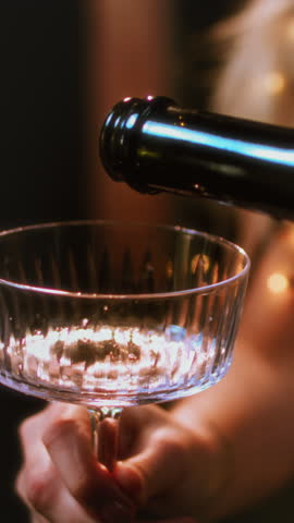 Vertical: girls pour champagne into round glasses from a bottle. Women in sparkling dress. Festive decoration, Christmas garland. Full cocktail creamers - le coupe. Female