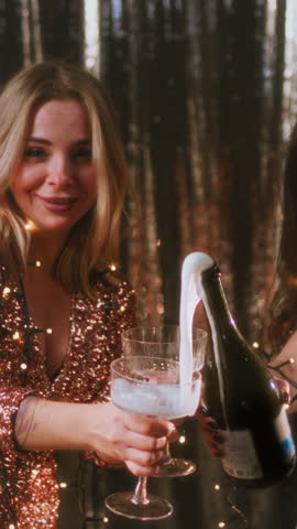 The girl opens a bottle of champagne. A woman in a sparkling dress uncorks a bottle of alcohol. White foam flows out of the neck of the bottle. The female is poured into glasses. Festive decoration