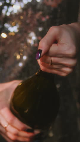 Vertical: girl opens a bottle of champagne. A woman in a sparkling dress uncorks a bottle of alcohol. White foam flows out of the neck of the bottle. Festive decoration, Christmas garland