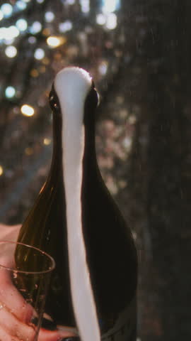 Vertical: girl opens a bottle of champagne. A woman in a sparkling dress uncorks a bottle of alcohol. White foam flows out of the neck of the bottle. Festive decoration, Christmas garland