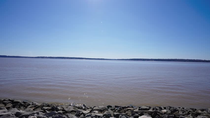 Wide view of the James River under a clear blue sky, with rocky shoreline in the foreground and distant tree-lined banks across the water