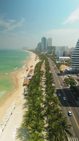 Aerial panorama revealing my khe beach coastline in da nang, vietnam, featuring lush palm trees, quiet roadway, modern urban skyline against pristine azure sky