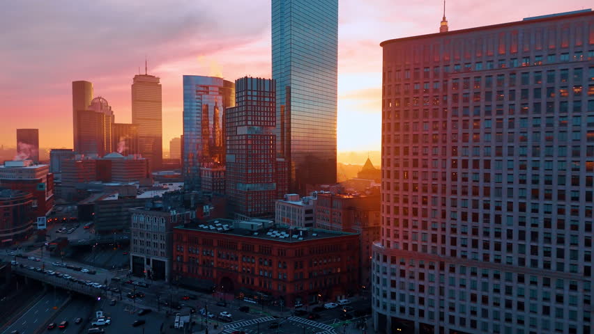 Setting sun lights the sky orange behind the high buildings of city downtown. Drone footage above Boston, Massachusetts, USA at sunset.