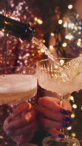The girls pour champagne into round glasses from a bottle. Women in a sparkling dress. Festive decoration, Christmas garland. Full cocktail creamers - le coupe. The female's hand holds a wine glass