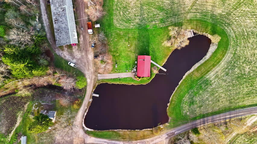 Overhead shot of a red-roofed house by a pond, surrounded by fields and dirt roads. Europe