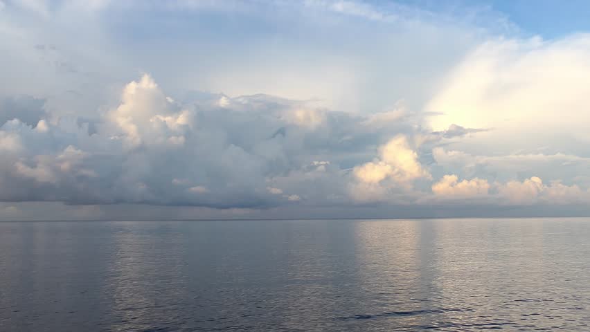 Majestic, sun-kissed clouds drift over a calm sea, their light and forms beautifully mirrored on the water