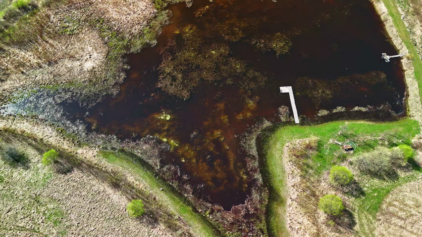 Drone footage of a dark, shallow pond surrounded by grassy banks and two wooden docks in the countryside