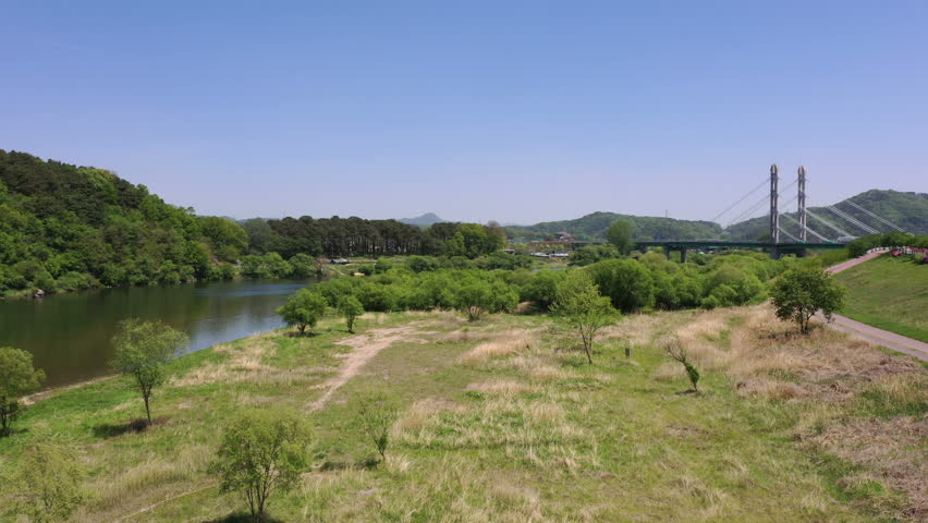 Aerial view of riverside park with walking and bike trail with bridge in background under clear blue sky.