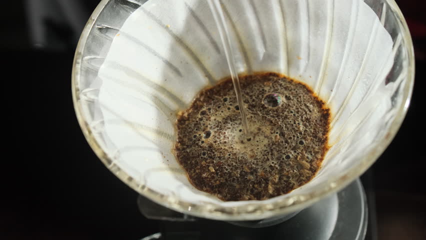 Person pours water into cloth coffee filter placed on carafe using teapot. Barista professionally controlls flow to create perfect cup of coffee