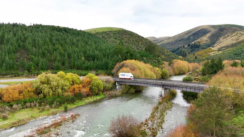 A vehicle crosses a bridge surrounded by lush greenery and hills under soft daylight at Lake Tekapo