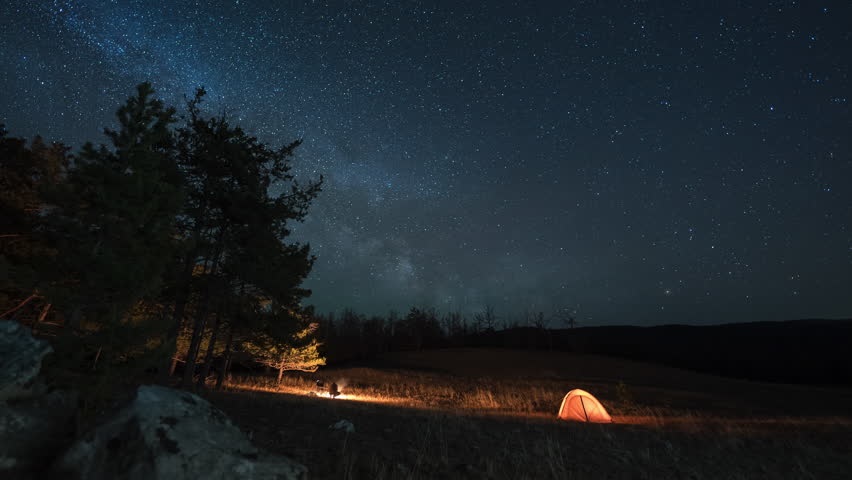 Night camping in mountains. Orange tent and campfire under moving Milky way
