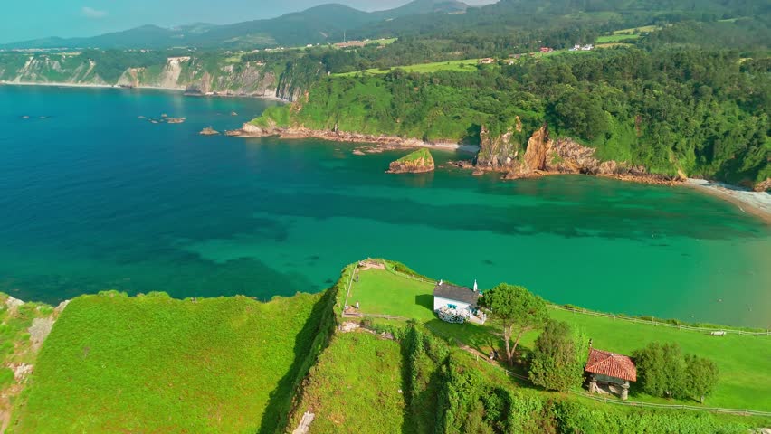 Aerial view of a small hermitage surrounded by stunning nature of Asturias in northern Spain. Ermita de la Regalina on the steep cliffs of the Cantabrian Sea coast. Beautiful Playa de Cadavedo beach