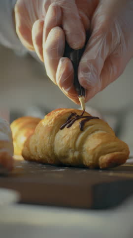 In a warm and inviting kitchen, a professional baker dressed in an apron carefully decorates a fresh croissant with chocolate. This delightful process showcases skilled pastry artistry