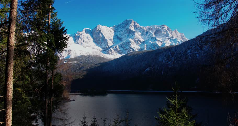 Majestic snow-capped mountains rise above serene lake surrounded by pine trees in winter landscape