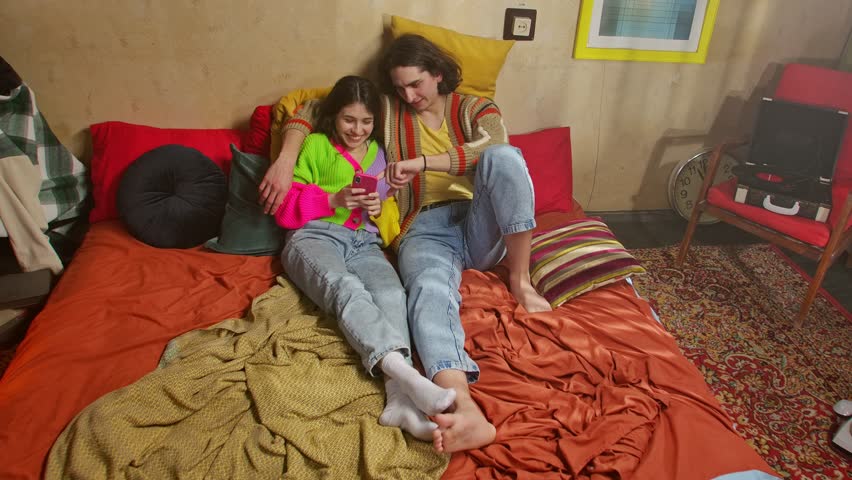 A joyful couple sits on a bed, sharing a moment and looking at a smartphone, surrounded by cozy, colorful decor.