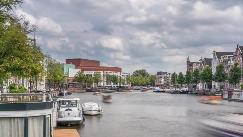 Hyperlapse of the exterior facade of the National Opera music and theatre venue. Houseboats on the river in Amsterdam under a cloudy sky timelapse. Popular tourist destination in the Netherlands