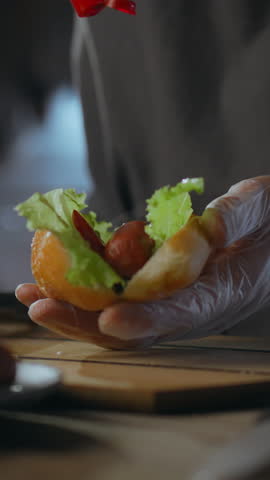 A skilled hot dog maker places a sausage into a fresh bun and evenly applies ketchup while adding lettuce. This culinary moment takes place in a food stall aimed at creating satisfying street food