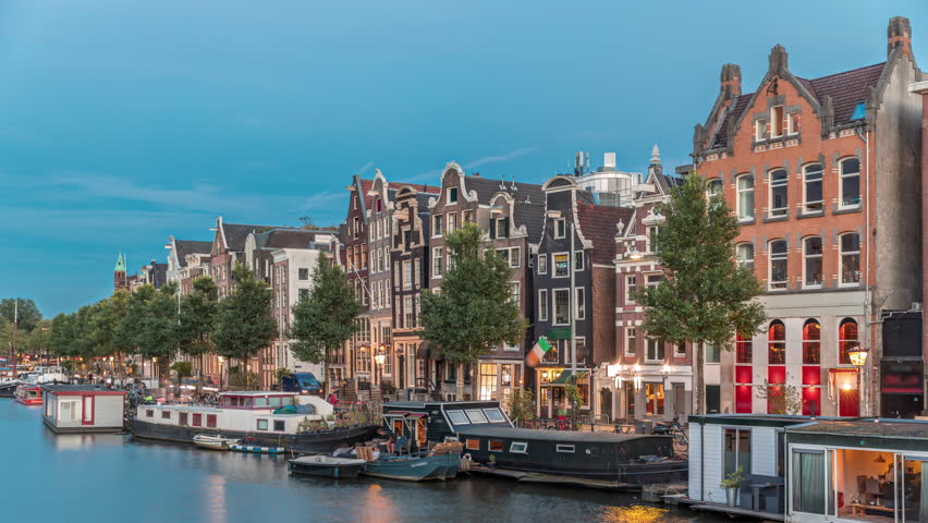 Panoramic timelapse hyperlapse of the illuminated famous Dancing Houses along the Amstel River day to night transition. Water channels with houseboats. Iconic landmark in Amsterdam, Netherlands