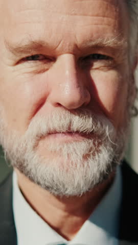 Vertical video, Portrait of a confident senior businessman with a white beard and long gray hair, standing in front of a modern office building.