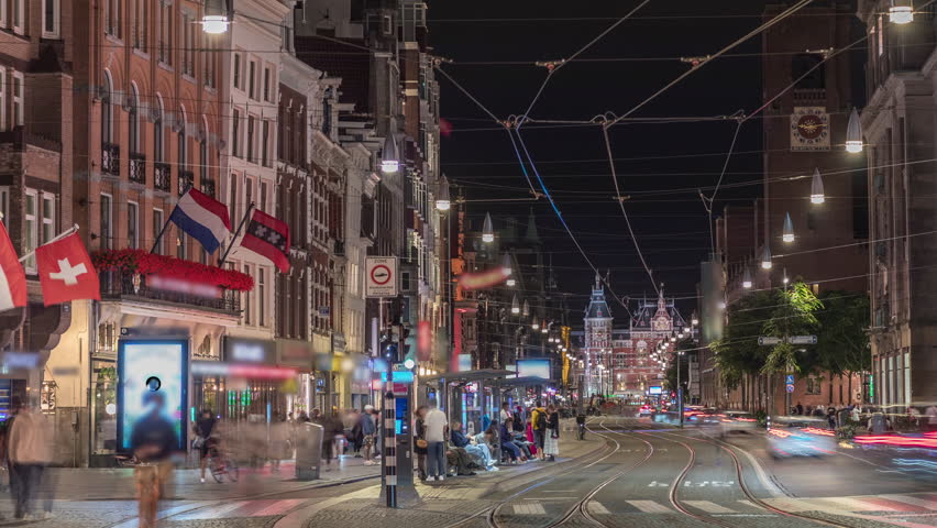 Electric tram on Damrak Street heading from Dam Square to Centraal Station in a timelapse at night. Traffic moves on the road while people wait at the tram stop. Amsterdam, Netherlands