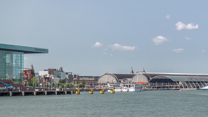 Panoramic view of Amsterdam Central Station with its curved roof and the Amstel River timelapse. A ferry crosses the river under a sunny sky with clouds vibrant movement. Amsterdam, Netherlands