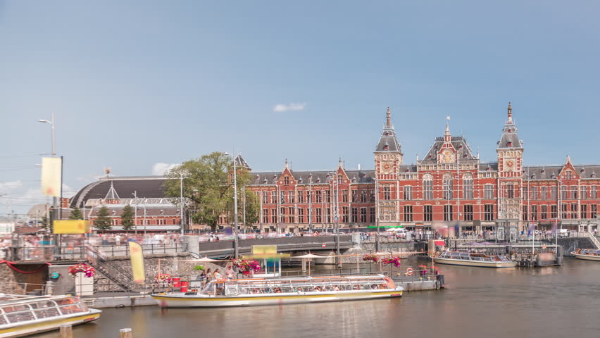 Timelapse of Amsterdam Central Railway Station front view with tour boats moored at the jetty, trams passing, and a canal in the foreground. Amsterdam, The Netherlands