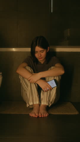 Full vertical of desperate young Caucasian girl sitting alone on floor in bathroom, clutching smartphone, crying, upset about relationship breakup, bullying or harassment